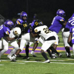 High school football players in purple and white uniforms compete under stadium lights during a nighttime game.