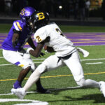 Two high school football players in purple and white uniforms collide on a field under stadium lights during a nighttime game.