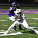 Two high school football players during a night game, one in a white uniform running with the ball as another in a purple uniform tries to tackle him.