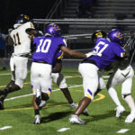 High school football players in purple and white uniforms block opponents in black and white during a night game on a turf field.