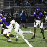 Two high school football teams, one in purple and one in white, are engaged in a tackle during a nighttime game on a turf field.