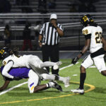 Football players in action as one tackles another to the ground, while a referee and a teammate observe on a lit field at night.