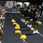Cheerleaders in black and gold uniforms perform on a track at night, with yellow pom-poms lined up in front of them and a crowd watching.
