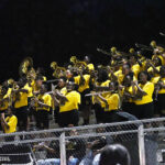 A marching band dressed in yellow shirts plays brass instruments in stadium bleachers at night, with a dark sky in the background.