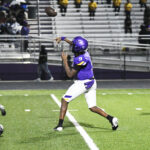 A football player in a purple uniform prepares to throw a pass on a lit field, with empty bleachers and spectators in the background.