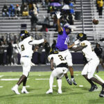 A football player in purple jumps to block a pass while three players in white uniforms defend on a field during a game.