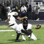 Football player in a white uniform kicks a field goal while a teammate holds the ball during a game on a green field.