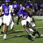 High school football players in purple and white uniforms compete during a nighttime game on a well-lit field. One player carries the ball while others attempt to tackle him.
