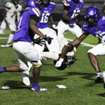 Football players in purple uniforms tackle an opposing player on a green field during a nighttime game.