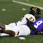 Football player in a white uniform tackles a player in a purple uniform on a green field during a game.
