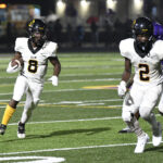 Two American football players in white uniforms running on a field at night, one carrying the ball, with stadium lights and blurred spectators in the background.