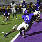 A football player in purple tackles an opponent near the end zone, while teammates and opponents watch the play on a brightly lit field.