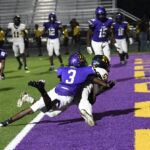 Football player in white dives into the end zone for a touchdown as a defender in purple attempts to tackle him, with other players watching in the background.