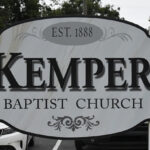 A sign reads "Kemper Baptist Church, Est. 1888," with decorative elements; trees and cars are visible in the background.