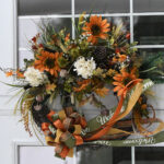Autumn wreath with orange and white flowers, greenery, pinecones, and a decorative ribbon bow, hanging on a glass door.