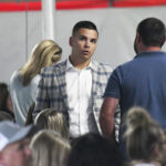 A man in a plaid jacket stands among a crowd, engaged in conversation with another man, while people sit and talk in the background.