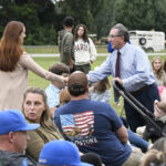 A man in a dress shirt and tie greets a woman in an outdoor gathering, with people sitting on grass and a trailer in the background.