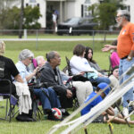 A group of people sit on lawn chairs outdoors, listening to a man in an orange shirt speaking, with houses and trees in the background.