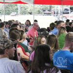 A large group of people seated outdoors under a tent, listening attentively, with trees and vehicles visible in the background.