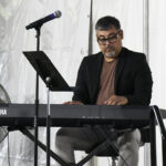 A man wearing glasses plays an electric keyboard on stage, with a microphone and sheet music stand nearby.