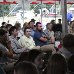 A group of people seated under a tent canopy, attentively watching or listening to an event. Some appear thoughtful while others face the front.