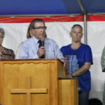 A man speaks into a microphone at a wooden podium with a cross, flanked by three people, under a tent with a white and red background.