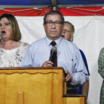 A man stands at a podium holding a microphone, with three women standing behind him, inside a tent with red, white, and blue decorations.