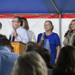 A group of people sing and speak on stage under a tent, addressing an audience seated before them.