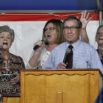 Four people stand behind a podium, singing or speaking into microphones at an event, with a red, white, and blue background.