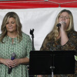 Two women stand on stage, one singing into a microphone and the other holding a microphone, with music stands and a red and white background behind them.