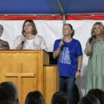 Four women stand on stage under a tent, holding microphones and singing behind a wooden podium with a cross, while an audience watches.