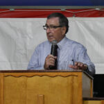 A man wearing glasses and a striped shirt speaks into a microphone while standing at a wooden podium.