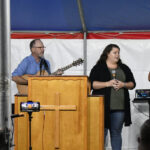 Man playing guitar and two women standing behind a wooden pulpit, speaking or singing, under a tent with microphones and recording equipment visible.
