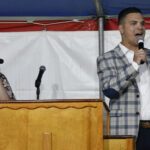 A man and a woman stand behind a wooden podium under a tent, both holding microphones and speaking to an audience.