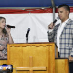 A woman and a man sing into microphones behind a wooden pulpit with a cross, under a tent with a red and white backdrop.