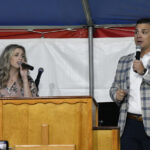 A woman and a man sing into microphones while standing behind a wooden podium under a tent with a red, white, and blue backdrop.