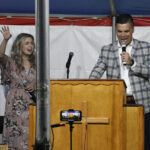 A man in a checked jacket speaks at a podium with a microphone, while a woman beside him smiles and raises her hand.