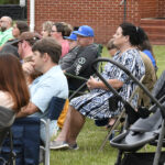A group of people sit on chairs outdoors, attentively watching an event. Some are dressed casually, and a stroller is visible in the foreground.