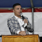 Man in plaid jacket speaking into a microphone while standing behind a wooden podium under a tent with a white and red background.