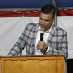 A man in a plaid jacket speaks into a microphone while standing at a wooden podium against a white and red backdrop.