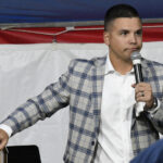 Man in plaid blazer holding microphone, standing in front of a red, white, and blue backdrop, speaking or presenting at an event.