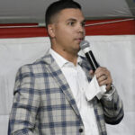 Man in plaid jacket holding a microphone and a piece of paper, speaking at an indoor event with a white background and red accent.