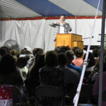 A speaker addresses a seated audience under a large tent, with people attentively listening in a dimly lit setting.