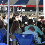 A group of people seated under a large tent, listening attentively to an event or speaker during the daytime.