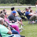 People sitting on folding chairs outdoors on grass, some with strollers and children, watching an event on a cloudy day.