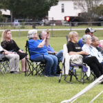 A group of people sitting on folding chairs outdoors, watching an event on a grassy field with houses and trees in the background.