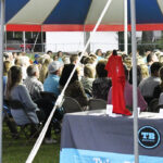 A large group of people sit under a blue and white tent, facing forward. A table with a red cloth and some items is in the foreground.