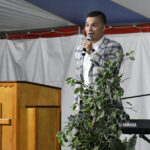A man in a plaid suit speaks into a microphone on stage, standing beside a wooden podium and a Yamaha keyboard, with a plant in the foreground.