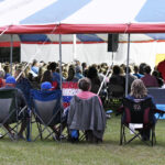 People sitting in folding chairs under a red, white, and blue tent at an outdoor event, listening to a speaker or performance.