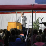 A man speaks at a podium under a tent to an audience, with a microphone, plants, and musical stands visible in the background.